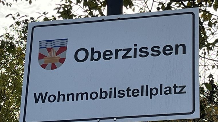 Ein Schild mit der Aufschrift „Oberzissen“ und „Wohnmobilstellplatz“. Es zeigt, dass dies ein ausgewiesener Platz für Wohnmobile ist.