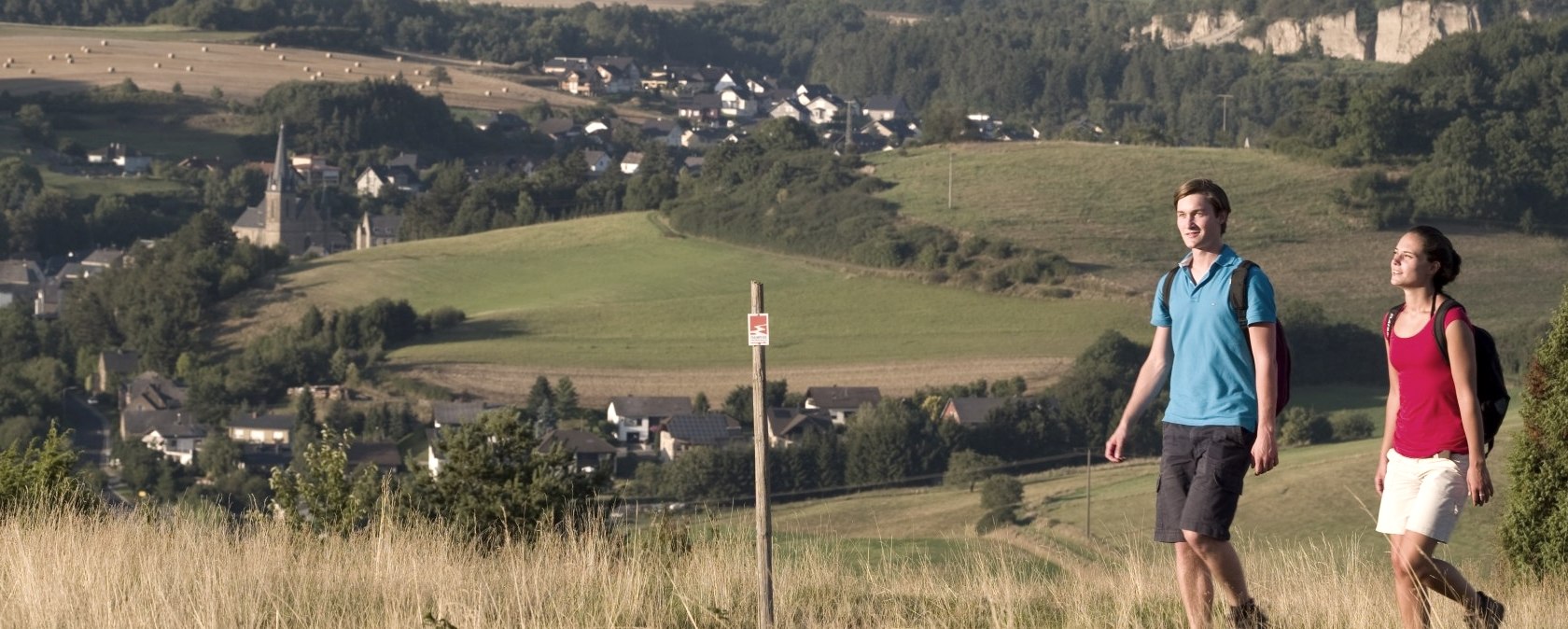Zwei Wanderer gehen durch eine hügelige Landschaft. Im Hintergrund sind ein Dorf und Felder zu sehen., © Traumpfade/Kappest