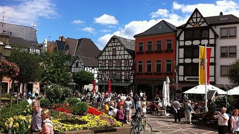 Ein lebhafter Marktplatz mit historischen Fachwerkhäusern und bunten Blumenbeeten. Viele Menschen genießen die Atmosphäre und die sonnigen Wetterverhältnisse.