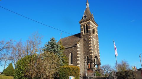 A charming church with a tall tower stands under a clear blue sky. Surrounding it are green trees and bushes.