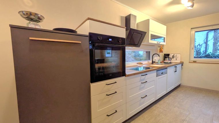 Eine moderne Küche mit weißen Schränken und einem Kühlschrank. Sie verfügt über einen Backofen und eine Spüle, sowie große Fenster, die natürliches Licht einlassen.