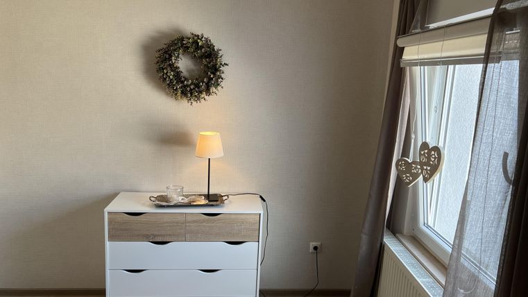 Ein gemütlicher Raum mit einem weißen Sideboard und einer Lampe. An der Wand hängt ein Kranz, und ein Fenster mit Vorhängen lässt Licht herein.