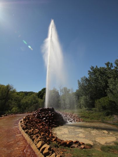 Ein Springbrunnen spritzt Wasser hoch in den klaren blauen Himmel. Umgeben von Bäumen und einem Naturweg wirkt die Szene erfrischend und einladend.
