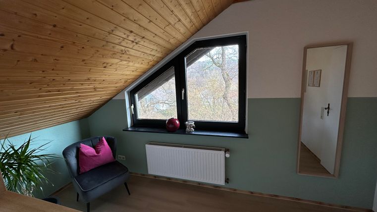 Ein gemütlicher Raum unter dem Dach mit einer Holzdecke. Es gibt ein Fenster mit Blick nach außen, einen Sessel und einen Spiegel.