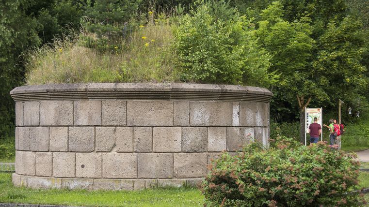 Ein runder Steinturm mit üppigem Grün auf der Spitze steht in einem Park. Im Hintergrund sind zwei Personen und ein Informationstafel zu sehen.