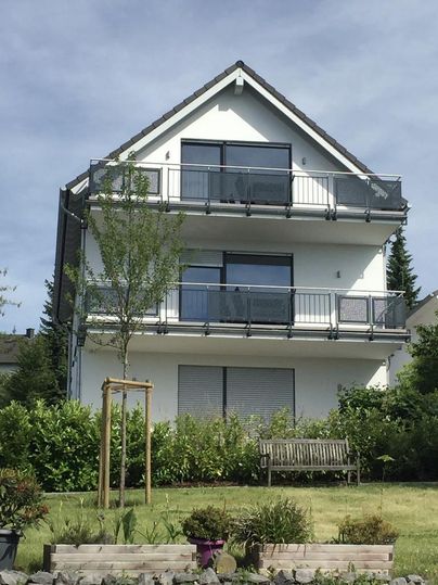 Ein modernes, dreistöckiges Haus mit Balkonen und großen Fenstern steht in einem grünen Garten. Im Vordergrund steht eine Holzbank und einige Pflanzen.