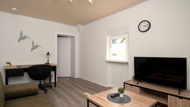 Ein modernes Wohnzimmer mit einem Sofa, einem Tisch und einem Fernseher. An der Wand hängen Bilder, und ein Schreibtisch mit einem Stuhl ist vorhanden.