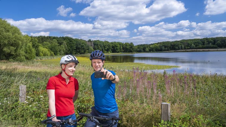 Zwei Radfahrer stehen lächelnd am Ufer eines Sees. Im Hintergrund sind grüne Bäume und ein blauer Himmel mit Wolken zu sehen.