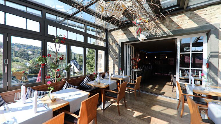 Ein lichtdurchfluteter Restaurantbereich mit Holzstühlen und eleganter Tischdekoration. Große Fenster bieten Blick ins Freie und sorgen für eine einladende Atmosphäre.