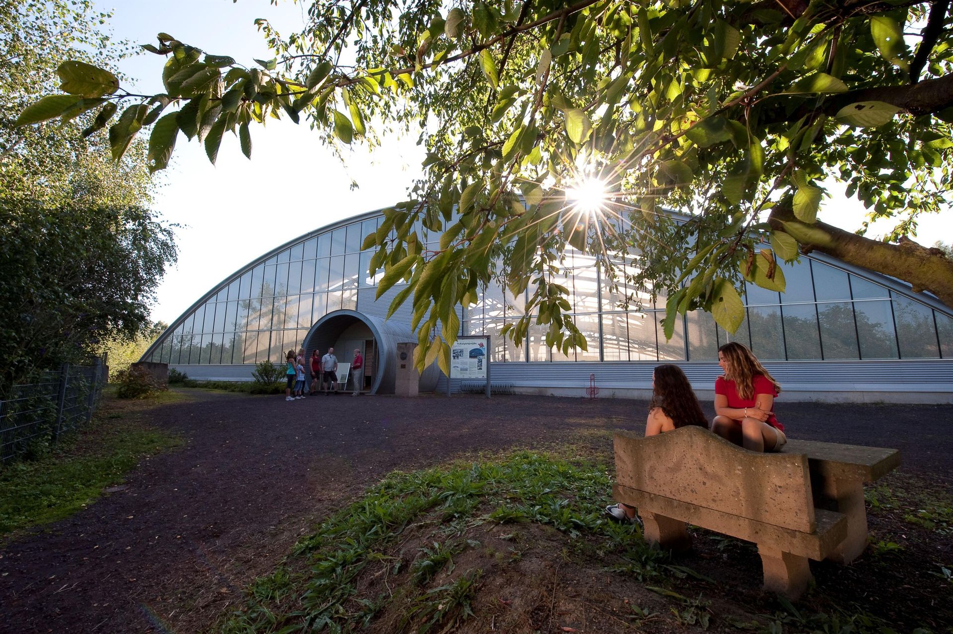 A modern greenhouse structure in nature. Two women are sitting on a bench and enjoying the sun.