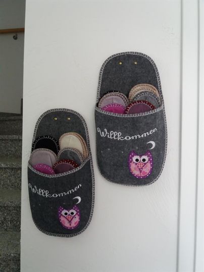 Zwei graue Filzhalter mit dem Schriftzug "Willkommen" hängen an der Wand. Sie sind mit bunten Hausschuhen gefüllt und zeigen eine niedliche Eule.