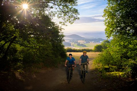 Zwei Fahrradfahrer auf einem Waldweg. Sie fahren Richtung Fotograf. Rechts und links von ihnen stehen grüne Bäume. Im Hintergrund eine hügelige Landschaft.