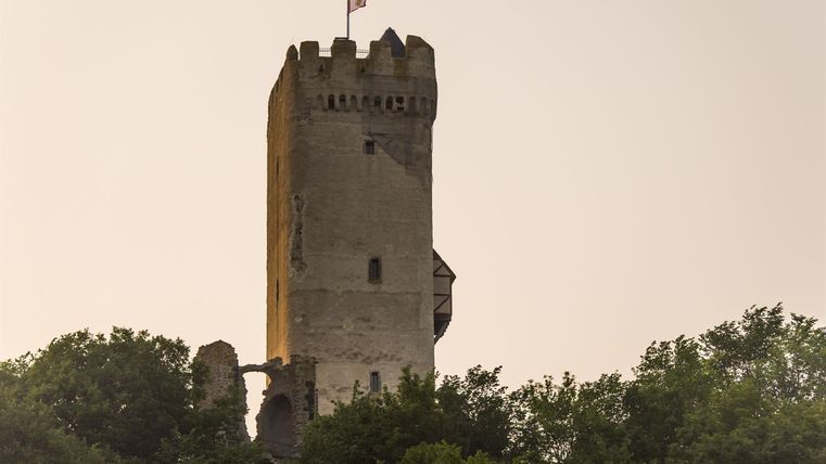 Ein alter Turm, umgeben von Bäumen, steigt in den Himmel. Eine Flagge weht oben auf dem Turm.