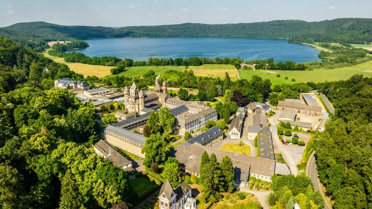 Eine beeindruckende Landschaft mit einem großen See, umgeben von grünen Wäldern und Wiesen. Im Vordergrund sind historische Gebäude zu sehen, die umgeben von schöner Natur liegen.