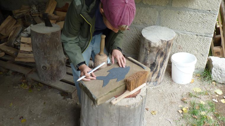 An older man and a boy are working together on a stone. The boy is wearing a mask and is watching attentively while the man shows him something.