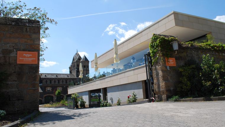 Ein modernes Gebäude mit großem Glasbereich und umliegendem Grün. Im Hintergrund sind historische Türme sichtbar.