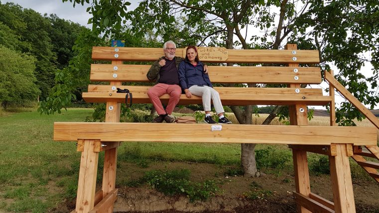 Ein großes Holzbänkchen steht in der Natur. Zwei Personen sitzen darauf und genießen die Umgebung.
