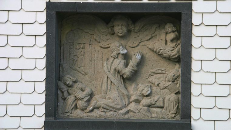 Ein historisches Relief mit religiösen Figuren. Es zeigt eine betende Person umgeben von anderen Gestalten und Engeln.