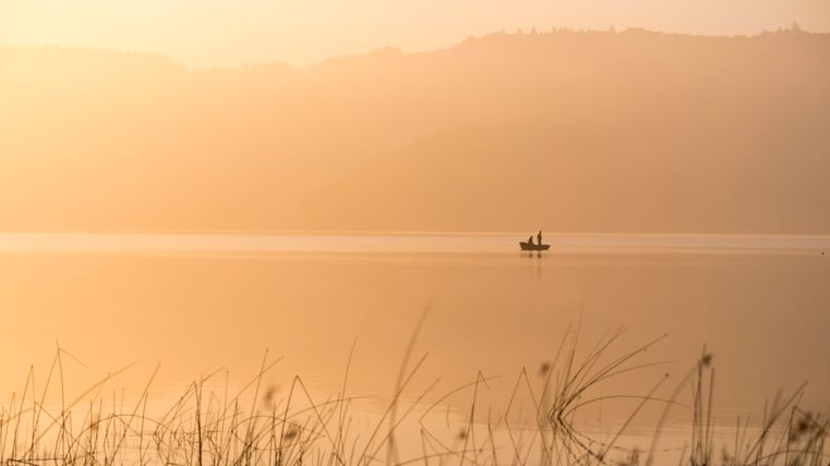 A tranquil lake with gentle hills in the background. A small boat glides across the water in a misty, golden light.