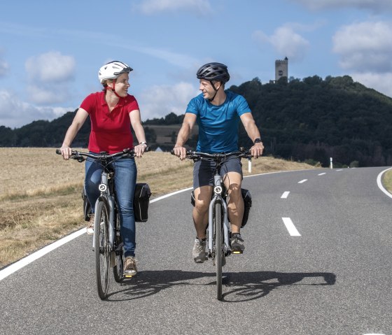 Zwei Fahrradfahrer fahren eine Stra&szlig;e entlang. Im Hintergrund die Burg Olbr&uuml;ck., &copy; Vulkanregion Laacher See, Klaus-Peter Kappest