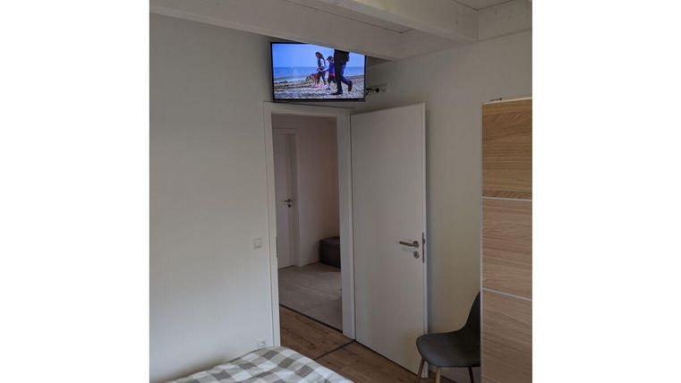 Ein gemütliches Schlafzimmer mit einem Fenster und einem kleinen Fernseher an der Wand. Das Bett hat eine kariertes Bettlaken und es gibt einen Stuhl in der Ecke.