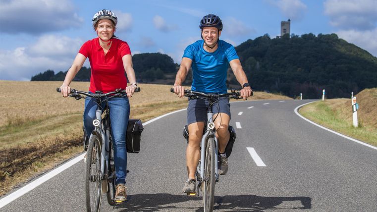 Zwei Radfahrer fahren auf einer Landstraße. Im Hintergrund ist ein sanfter Hügel und ein blauer Himmel zu sehen.