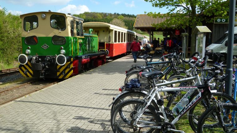 Eine kleine Lokomotive steht an einem Bahngleis, während Passagiere aussteigen. Neben der Lok sind zahlreiche Fahrräder abgestellt.