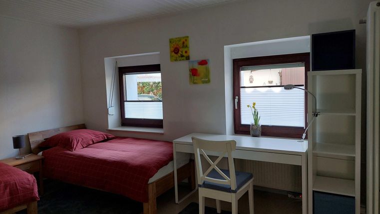 A cozy room with two beds, a desk, and a shelf. Bright walls and windows provide light and a friendly atmosphere.