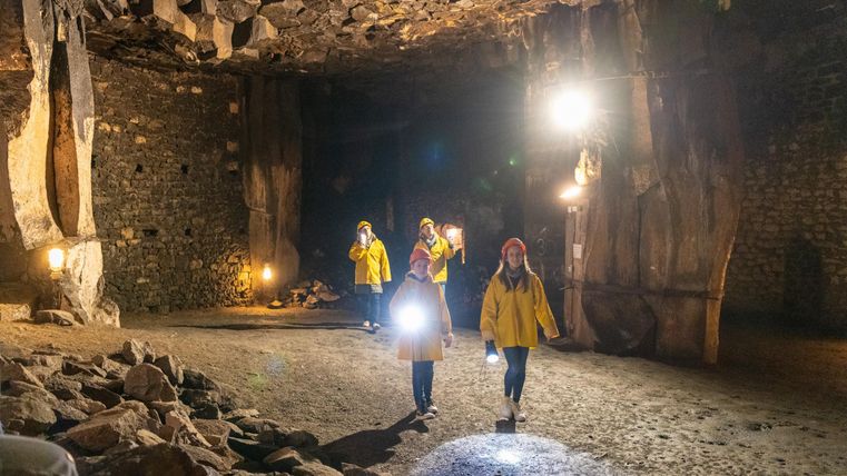 A group of people in yellow rain jackets explores a lit cave. They are holding flashlights and moving across the stone floor.