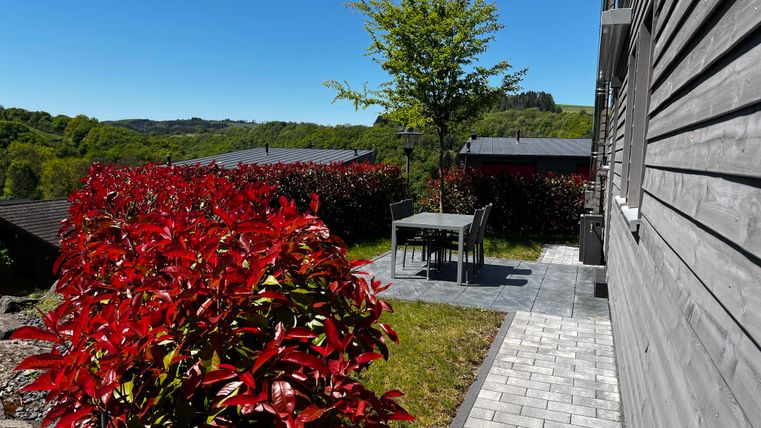 Ein ruhiger Garten mit bunten roten Pflanzen und einem grauen Tischset. Im Hintergrund sind grüne Hügel und eine klare blaue Himmelssicht sichtbar.