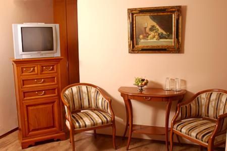Ein gemütlicher Raum mit einem Fernseher, einer Kommode und zwei Stühlen. An der Wand hängt ein schönes Gemälde und auf dem Tisch stehen Glaswasser und eine Fruchtbowl.