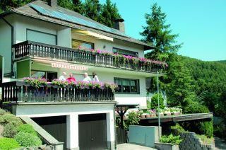 Ein modernes Haus mit einem Balkon, geschmückt mit bunten Blumen. Im Hintergrund sind grüne Hügel und Bäume zu sehen.