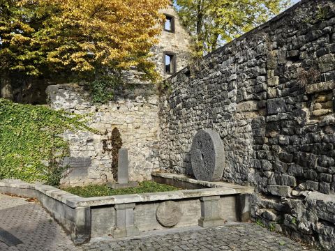 Ein historischer Platz mit einer alten Mauer und einer Steinmühle. Herbstliche Bäume im Hintergrund verleihen der Szene Farbe.
