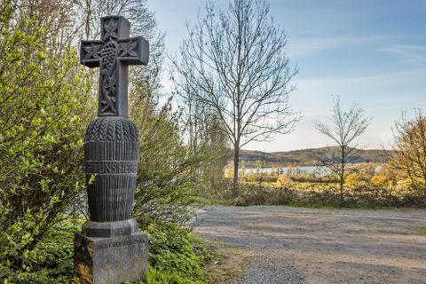 Ein kunstvoll geschnitztes Kreuz steht an einem Weg. Im Hintergrund erstreckt sich eine ruhige Landschaft mit Bäumen und Wasser.
