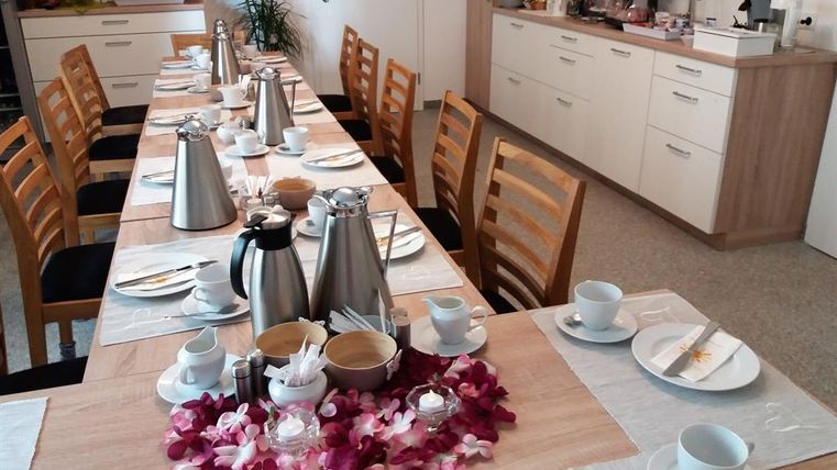 Ein gedeckter Tisch mit mehreren Kaffeekannen und Tassen, umgeben von Stühlen. Im Hintergrund ist eine Küchenzeile mit verschiedenen Leckereien sichtbar.