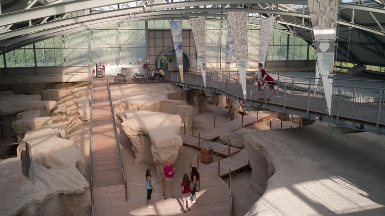 Eine archäologische Stätte mit ausgehobenen Strukturen in einem modernen Gebäude. Besucher erkunden den Raum auf Holzplanken und genießen die Lichtverhältnisse.