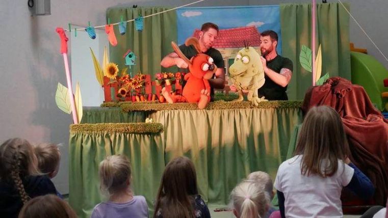 Ein Puppenspiel mit zwei Puppenspielern, die lebendige Figuren präsentieren. Im Hintergrund sind bunte Dekorationen und eine kleine Kulisse zu sehen.