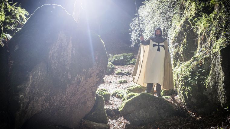 A knight stands in a forest, surrounded by large stones and moss-covered ground. He holds a sword and wears a cloak with a cross.