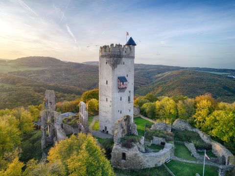 Eine beeindruckende Burgruine auf einem Hügel, umgeben von herbstlichen Bäumen. Der klare Himmel und die sanften Hügel im Hintergrund ergänzen die malerische Szene.