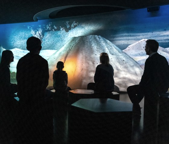 Besucher sitzen im Dunkeln vor einer beleuchteten Vulkanprojektion im Lava-Dome Mendig, die eine spannende Atmosph&auml;re schafft., &copy; Eifel Tourismus GmbH, Dominik Ketz