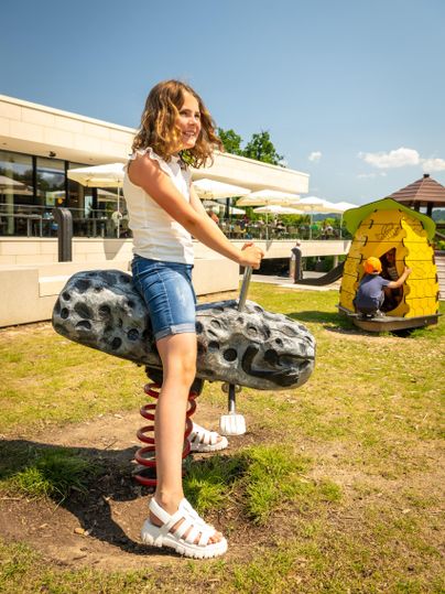 Ein Mädchen sitzt auf einem Federspielgerät im Freien. Im Hintergrund sind ein Spielhaus und das Gebäude einer Einrichtung zu sehen.