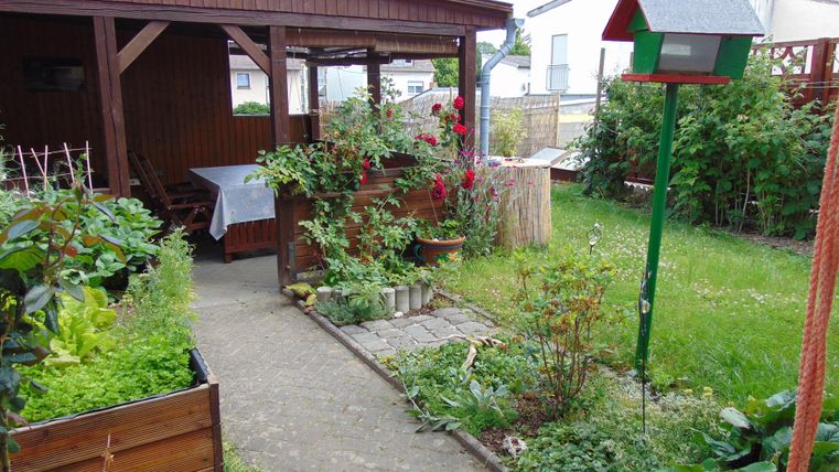 A cozy garden with a paved path, colorful plants, and a covered seating area. In the background, there are more houses and a birdhouse.