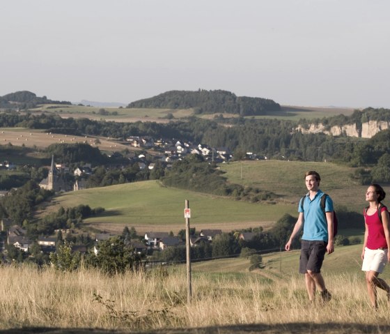 Zwei Wanderer in sommerlicher Kleidung gehen durch eine h&uuml;gelige Landschaft mit D&ouml;rfern und Feldern im Hintergrund., &copy; Traumpfade/Kappest