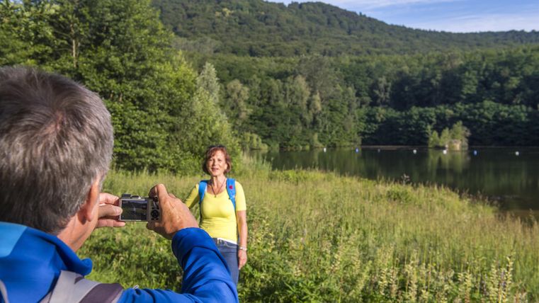 Eine Frau steht lächelnd vor einem malerischen See, während ein Mann sie mit einer Kamera fotografiert. Im Hintergrund sind grüne Hügel und strahlend blauer Himmel zu sehen.