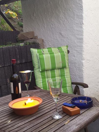 Ein gemütlicher Tisch auf einer Terrasse mit einem grünen Stuhl, einer Flasche Wein und zwei Gläsern. Ein brennendes Kerzenlicht sorgt für eine entspannte Atmosphäre.