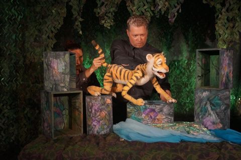 Ein Puppenspieler führt eine Tiger-Puppe auf einer bühnenähnlichen Fläche vor. Im Hintergrund sind grüne Pflanzen und dekorative Boxen zu sehen.