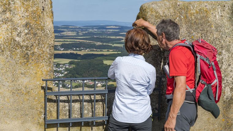 A couple stands by a wall and looks out at the landscape. In the background, gentle hills and a clear sky can be seen.