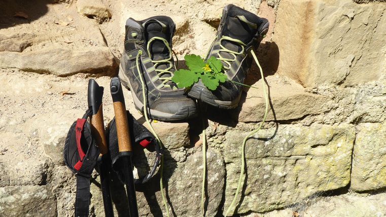 Hiking shoes are lying on a stone floor, surrounded by nature. Next to the shoes are a backpack and a hiking pole.