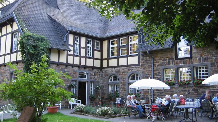 Ein hübsches Haus im Fachwerkstil mit einem Garten. Einige Menschen sitzen entspannt im Freien unter Sonnenschirmen.
