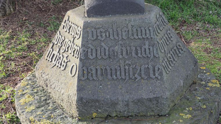 Ein steinerner Obelisk mit eingravierten Inschriften. Die Umgebung ist grün und naturalistisch.